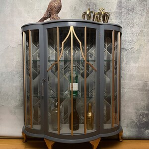 May include: A grey glass-fronted cabinet with gold accents and decorative feet. The cabinet has two doors and shelves inside. The cabinet is decorated with a geometric pattern and has a gold trim around the edges. The cabinet is sitting on a wooden floor.