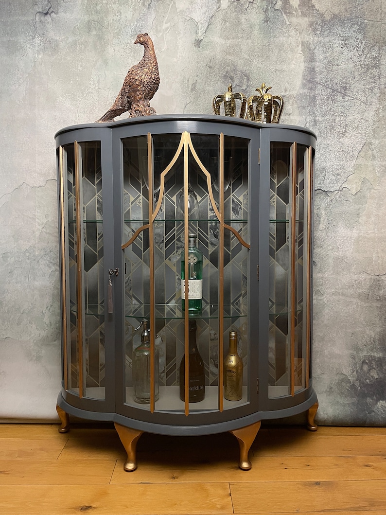 May include: A grey glass-fronted cabinet with gold accents and decorative feet. The cabinet has two doors and shelves inside. The cabinet is decorated with a geometric pattern and has a gold trim around the edges. The cabinet is sitting on a wooden floor.