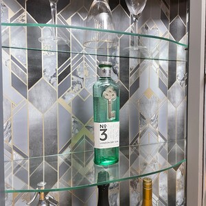 May include: A glass cabinet with shelves filled with various bottles and glasses. The back of the cabinet is lined with a geometric patterned wallpaper in shades of grey and gold.