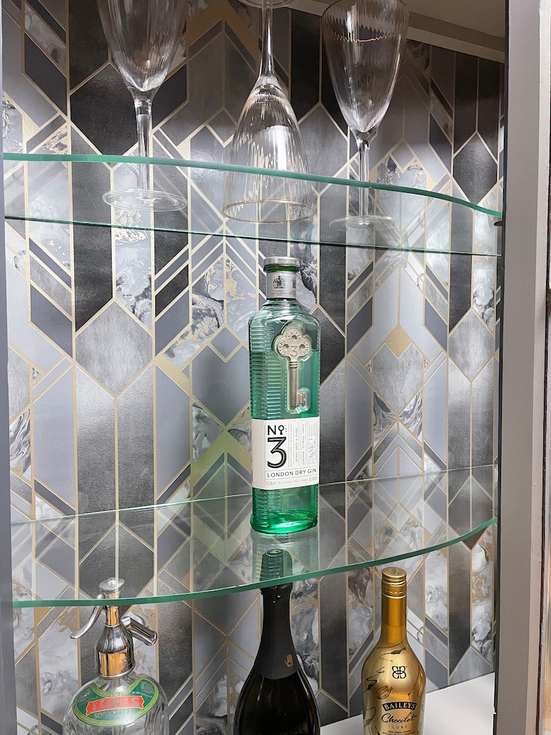 May include: A glass cabinet with shelves filled with various bottles and glasses. The back of the cabinet is lined with a geometric patterned wallpaper in shades of grey and gold.