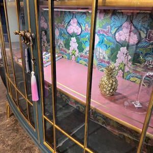 Puede incluir: Un armario de cristal azul oscuro con ribetes dorados y un interior rosa. El armario tiene una pi&ntilde;a y un vaso en el estante. El armario tiene un papel tapiz con estampado floral en la parte posterior.
