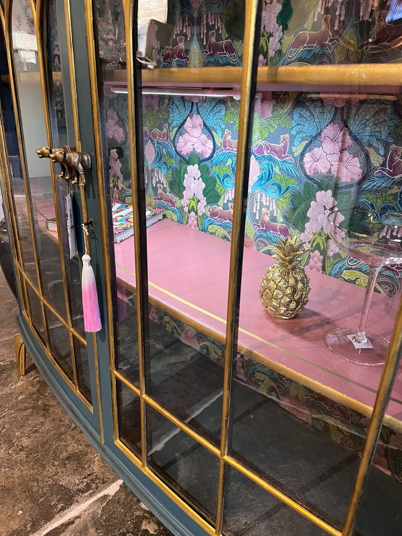 Puede incluir: Un armario de cristal azul oscuro con ribetes dorados y un interior rosa. El armario tiene una pi&ntilde;a y un vaso en el estante. El armario tiene un papel tapiz con estampado floral en la parte posterior.