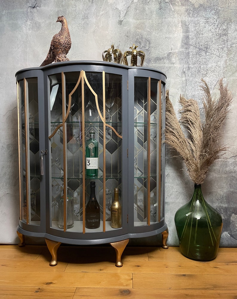 May include: A grey curved glass-fronted cabinet with gold accents and gold feet. The cabinet has two shelves and is filled with bottles and glasses. The cabinet is sitting on a wooden floor. There are two gold crowns on top of the cabinet and a pheasant figurine on the left side.