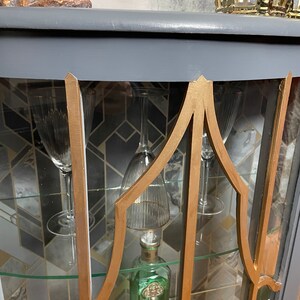 May include: A grey glass-fronted cabinet with gold trim and shelves. The cabinet has a curved top and is decorated with a geometric pattern. The shelves are filled with glassware and a bottle of gin.