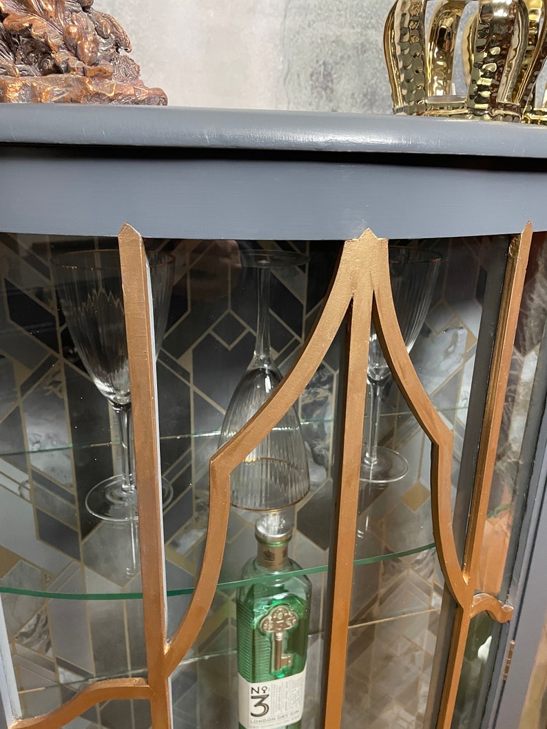 May include: A grey glass-fronted cabinet with gold trim and shelves. The cabinet has a curved top and is decorated with a geometric pattern. The shelves are filled with glassware and a bottle of gin.