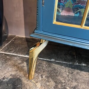 Puede incluir: Un armario azul verdoso con molduras doradas y paneles de vidrio decorativos. El armario tiene cuatro patas de metal dorado con un dise&ntilde;o decorativo.