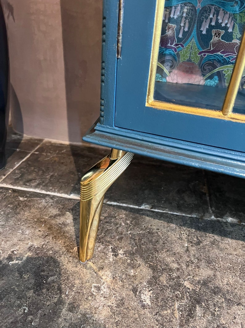 Puede incluir: Un armario azul verdoso con molduras doradas y paneles de vidrio decorativos. El armario tiene cuatro patas de metal dorado con un dise&ntilde;o decorativo.