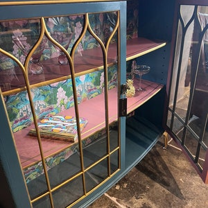 Puede incluir: Un armario azul verdoso con ribetes dorados y puertas de cristal. El armario tiene dos estantes y est&aacute; decorado con un papel tapiz floral rosa y verde. Un libro est&aacute; en el estante inferior.