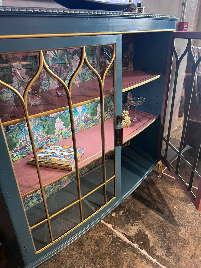Puede incluir: Un armario azul verdoso con ribetes dorados y puertas de cristal. El armario tiene dos estantes y est&aacute; decorado con un papel tapiz floral rosa y verde. Un libro est&aacute; en el estante inferior.