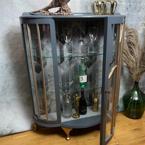 May include: A grey curved glass-fronted cabinet with gold accents and glass shelves. The cabinet has a decorative gold trim and is sitting on four gold feet. The cabinet is open and there are bottles and glasses inside.