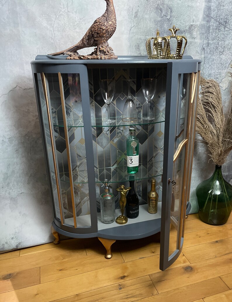 May include: A grey curved glass-fronted cabinet with gold accents and glass shelves. The cabinet has a decorative gold trim and is sitting on four gold feet. The cabinet is open and there are bottles and glasses inside.