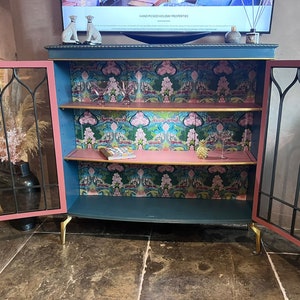 Puede incluir: Un armario azul verdoso con dos puertas de vidrio y dos estantes. El armario tiene un interior rosa y molduras doradas. Los estantes est&aacute;n forrados con un papel tapiz estampado con dise&ntilde;os florales rosas y verdes. El armario est&aacute; sobre patas doradas.
