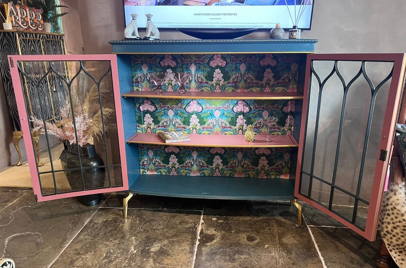 Puede incluir: Un armario azul verdoso con dos puertas de vidrio y dos estantes. El armario tiene un interior rosa y molduras doradas. Los estantes est&aacute;n forrados con un papel tapiz estampado con dise&ntilde;os florales rosas y verdes. El armario est&aacute; sobre patas doradas.