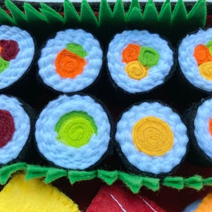 Puede incluir: Un conjunto de ocho rollos de sushi de fieltro con diferentes rellenos, incluyendo aguacate, pepino y cangrejo de imitaci&oacute;n. Los rollos est&aacute;n dispuestos en una fila sobre un fondo de fieltro rojo y verde.