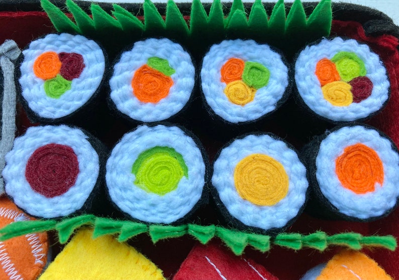Puede incluir: Un conjunto de ocho rollos de sushi de fieltro con diferentes rellenos, incluyendo aguacate, pepino y cangrejo de imitaci&oacute;n. Los rollos est&aacute;n dispuestos en una fila sobre un fondo de fieltro rojo y verde.