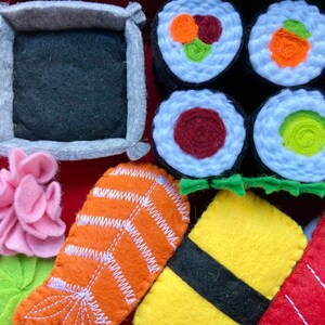 Puede incluir: Conjunto de comida de sushi de fieltro con varios rollos de sushi, una pieza de salm&oacute;n, un cuadrado amarillo de arroz y una flor rosa. Los rollos de sushi est&aacute;n hechos de fieltro blanco con rellenos de diferentes colores. El salm&oacute;n es naranja con rayas blancas. El arroz es amarillo con una tira negra de alga. La flor es rosa con un tallo verde.