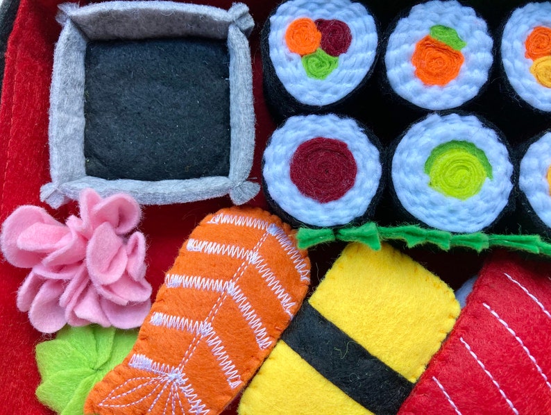 Puede incluir: Conjunto de comida de sushi de fieltro con varios rollos de sushi, una pieza de salm&oacute;n, un cuadrado amarillo de arroz y una flor rosa. Los rollos de sushi est&aacute;n hechos de fieltro blanco con rellenos de diferentes colores. El salm&oacute;n es naranja con rayas blancas. El arroz es amarillo con una tira negra de alga. La flor es rosa con un tallo verde.