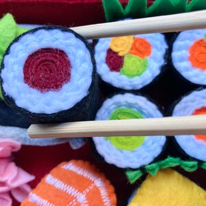 A set of felt sushi rolls hold with chopsticks to demonstrate the size. The rolls are made of felt and have different fillings, including avocado, salmon, and tuna. The rolls are arranged on a red felt surface which is the bottom of the bento box.
