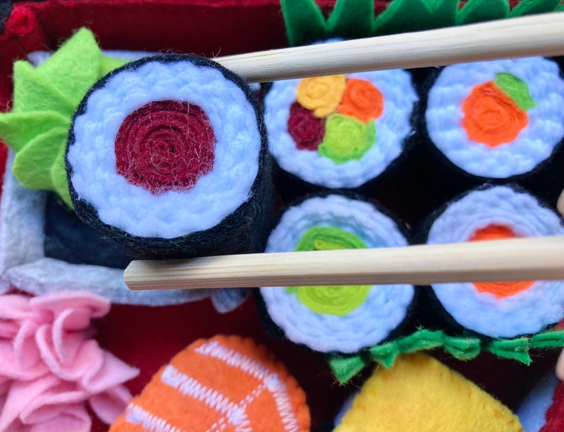A set of felt sushi rolls hold with chopsticks to demonstrate the size. The rolls are made of felt and have different fillings, including avocado, salmon, and tuna. The rolls are arranged on a red felt surface which is the bottom of the bento box.