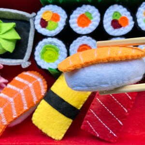 Puede incluir: Un conjunto de sushi de fieltro, que incluye nigiri, maki y sashimi, con palillos. El sushi est&aacute; hecho de fieltro y tiene detalles realistas, como semillas de s&eacute;samo y algas.