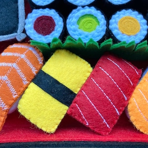 Puede incluir: Un conjunto de rollos de sushi de fieltro, que incluyen salm&oacute;n, at&uacute;n, pez lim&oacute;n y aguacate. El sushi est&aacute; dispuesto sobre una estera de fieltro roja con un fondo de fieltro negro.