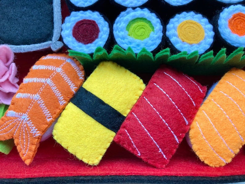 Puede incluir: Un conjunto de rollos de sushi de fieltro, que incluyen salm&oacute;n, at&uacute;n, pez lim&oacute;n y aguacate. El sushi est&aacute; dispuesto sobre una estera de fieltro roja con un fondo de fieltro negro.