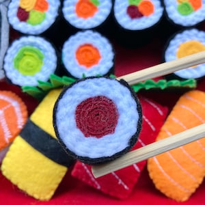 Puede incluir: Primer plano de un rollo de sushi de fieltro con relleno rojo y blanco. El rollo de sushi est&aacute; siendo sostenido por palillos sobre un plato de otros rollos de sushi y sashimi de fieltro.