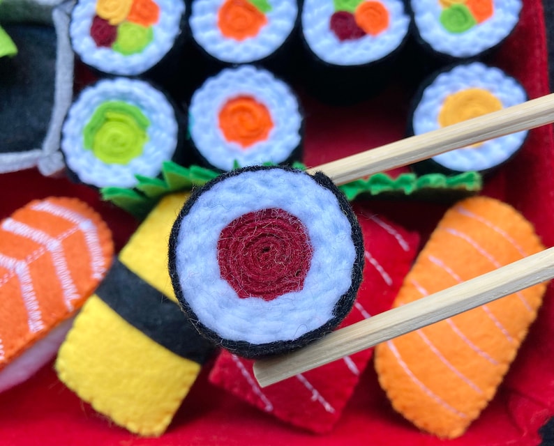 Puede incluir: Primer plano de un rollo de sushi de fieltro con relleno rojo y blanco. El rollo de sushi est&aacute; siendo sostenido por palillos sobre un plato de otros rollos de sushi y sashimi de fieltro.