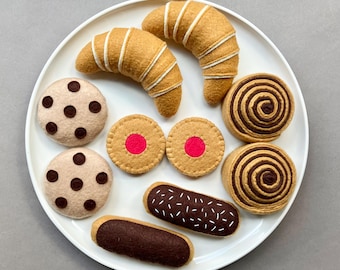 Puede incluir: Un plato blanco con una variedad de alimentos de fieltro, incluyendo croissants, galletas y pasteles. Las galletas están decoradas con chispas de chocolate y los pasteles tienen un diseño de espiral.