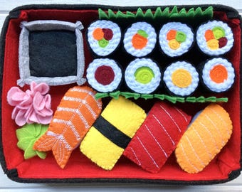 Puede incluir: Un juego de sushi de fieltro en una caja bento roja y negra. El juego incluye varios tipos de sushi, como nigiri, maki y sashimi, así como un pequeño recipiente para salsa de soja.