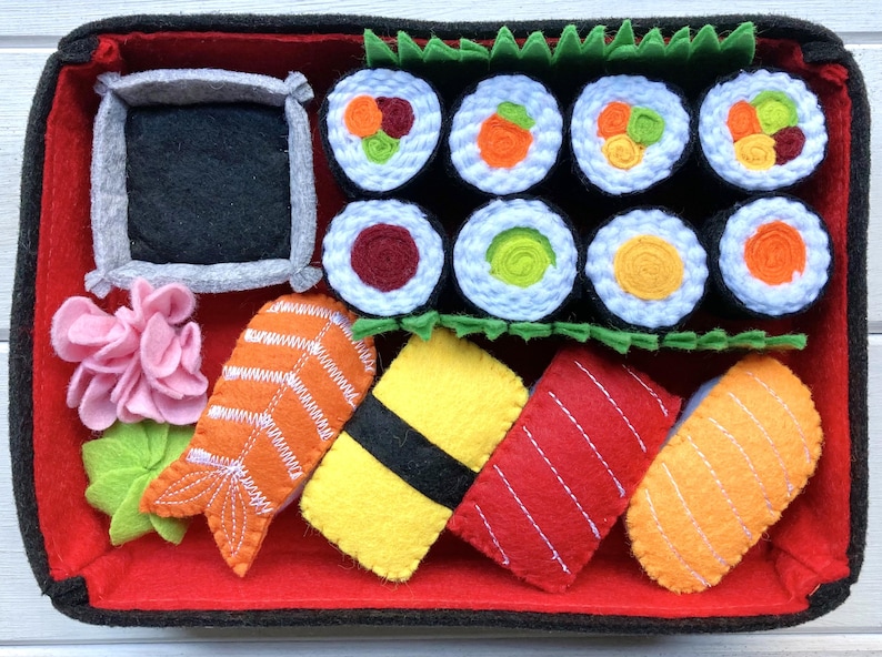 A handmade felt sushi play set in a red and black bento box. The set includes various types of sushi, including nigiri, maki, and sashimi, as well as a small container for soy sauce.