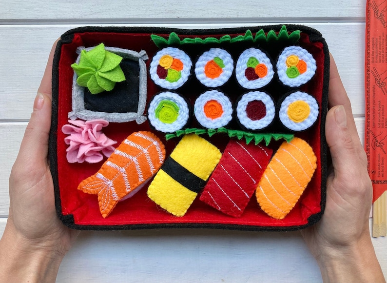 A red felt sushi tray with a variety of felt sushi pieces, including salmon, tuna, yellowtail, and cucumber rolls. The tray also includes a small dish of wasabi and a piece of pickled ginger. The bento box that contain all the items are held to demonstrate the size of the set