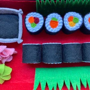Puede incluir: Un juego de sushi de fieltro con cuatro piezas de sushi, cuatro rollos de sushi, un taz&oacute;n peque&ntilde;o, una flor rosa y una hoja verde. El sushi est&aacute; decorado con verduras de fieltro.