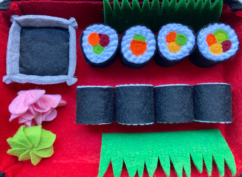 Puede incluir: Un juego de sushi de fieltro con cuatro piezas de sushi, cuatro rollos de sushi, un taz&oacute;n peque&ntilde;o, una flor rosa y una hoja verde. El sushi est&aacute; decorado con verduras de fieltro.
