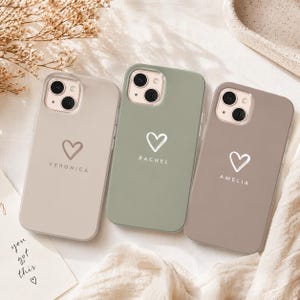 May include: Three phone cases in neutral colors: beige, sage green, and taupe. Each case features a white heart graphic and a name: Veronica, Rachel, and Amelia. The cases are on a white surface with a handwritten note that says "you got this".