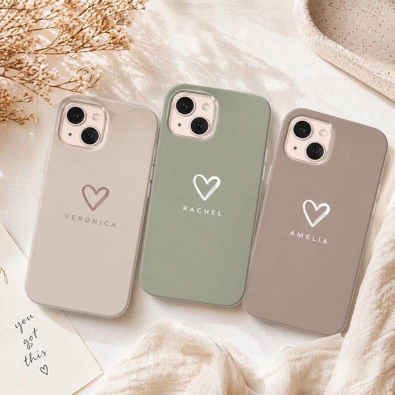 May include: Three phone cases in neutral colors: beige, sage green, and taupe. Each case features a white heart graphic and a name: Veronica, Rachel, and Amelia. The cases are on a white surface with a handwritten note that says "you got this".