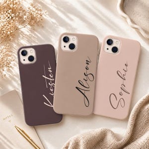 May include: Three personalized phone cases in shades of brown, tan, and pink. Each case has a name written in a cursive font. The names are Kirsten, Alison, and Sophie. The cases are on a white surface with a notebook and pen.