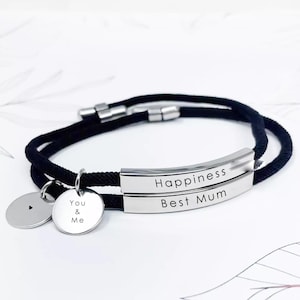 Może przedstawiać: Bransoletka z czarnego sznurka z dwoma srebrnymi paskami grawerowanymi "Happiness" i "Best Mum". Dołączony jest mały srebrny zawieszek z napisem "You & Me".