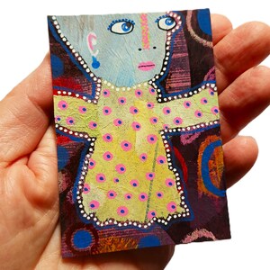 May include: A small rectangular painting on paper featuring a stylized figure with a light blue face, blue eyes, and pink lips. The figure wears a yellow dress with pink polka dots and white dotted outlines, set against a dark purple background.