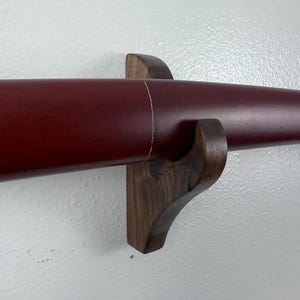 May include: A dark wood wall mount with a red katana sword scabbard resting on it. The scabbard has a gold accent on the front.