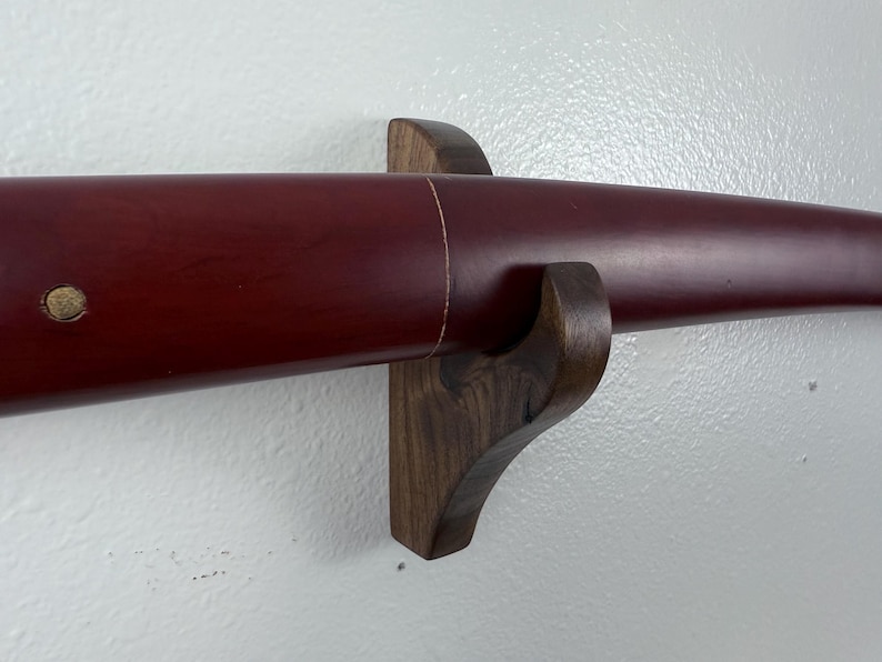 May include: A dark wood wall mount with a red katana sword scabbard resting on it. The scabbard has a gold accent on the front.