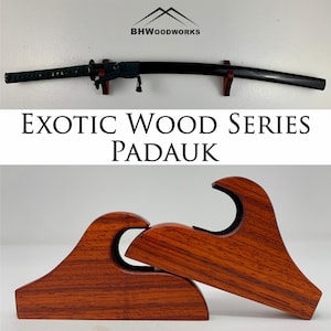 May include: A wooden wall mount for a katana sword, made from exotic padauk wood. The mount is shaped like a wave and has a dark brown color with a reddish hue. The text "Exotic Wood Series Padauk" is displayed on the image.