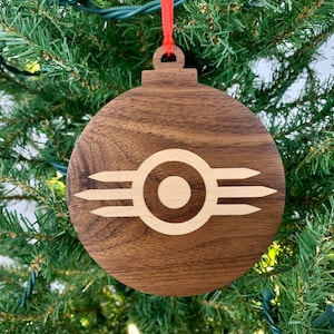 May include: A wooden Christmas ornament with a brown finish and a light-colored logo. The logo is a circle with three lines on each side. The ornament hangs from a red ribbon and is displayed on a Christmas tree.