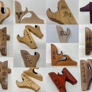 May include: A collection of 12 wooden wall-mounted sword holders with various Japanese kanji characters engraved on them. The holders are made of different types of wood, including light and dark wood, and some are stained in a deep purple color.