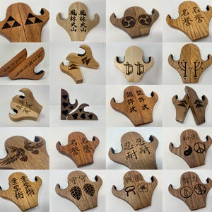 May include: A collection of 20 wooden guitar stands with various designs and engravings. The stands are made of light and dark wood and feature different patterns, symbols, and text. Some of the engravings include Japanese characters, a yin yang symbol, a peace sign, and a wolf's head.
