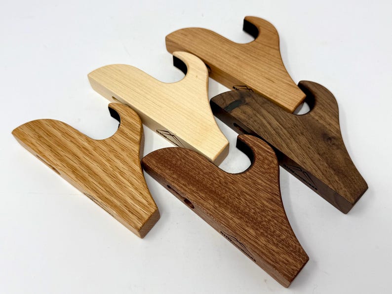 May include: Four wooden gun wall mounts in different wood types: maple, oak, walnut, and cherry. Each mount has a curved design and a black rubber insert for holding the gun.