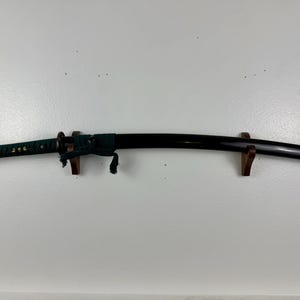 May include: A black katana sword with a green and gold wrapped handle, mounted on a wooden stand.