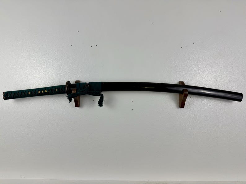 May include: A black katana sword with a green and gold wrapped handle, mounted on a wooden stand.