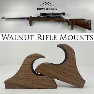 May include: Two walnut wood rifle mounts, each with a curved shape and a black rubber lining. The mounts are designed to hold a rifle securely on a wall.