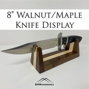 Puede incluir: Un expositor de cuchillos de nogal y arce, que sostiene un cuchillo grande. El cuchillo tiene una hoja plateada y un mango negro. El soporte de exhibición presenta una franja de madera clara. El texto dice: "8" Walnut/Maple Knife Display".